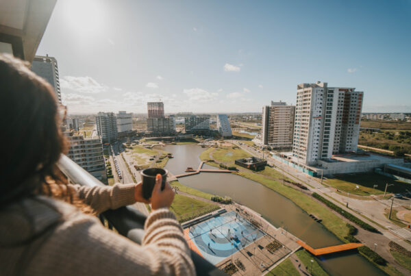Vista em perspectiva de uma mulher de costas, segurando uma caneca escura em uma sacada de andar alto. Ela observa um bairro planejado e ensolarado, que conta com prédios modernos, uma quadra poliesportiva nas cores azul e laranja, e um lago artificial sinuoso que corta a área verde. O céu está limpo e azul.